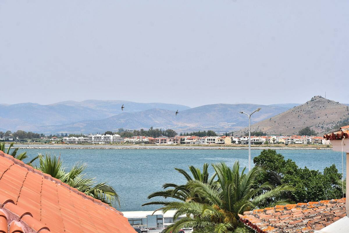 A coastal town with a view of the sea, mountains in the background, and a hill with a structure on top.