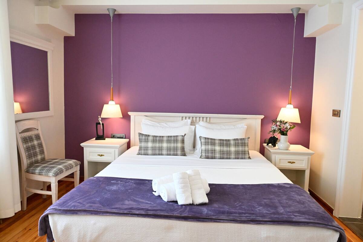 A bedroom with a purple wall, a bed with white bedding, two nightstands with lamps, a chair, and a window.
