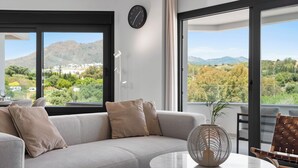 Een moderne woonkamer met een grijze bank, een ronde tafel en uitzicht op bergen en groen.