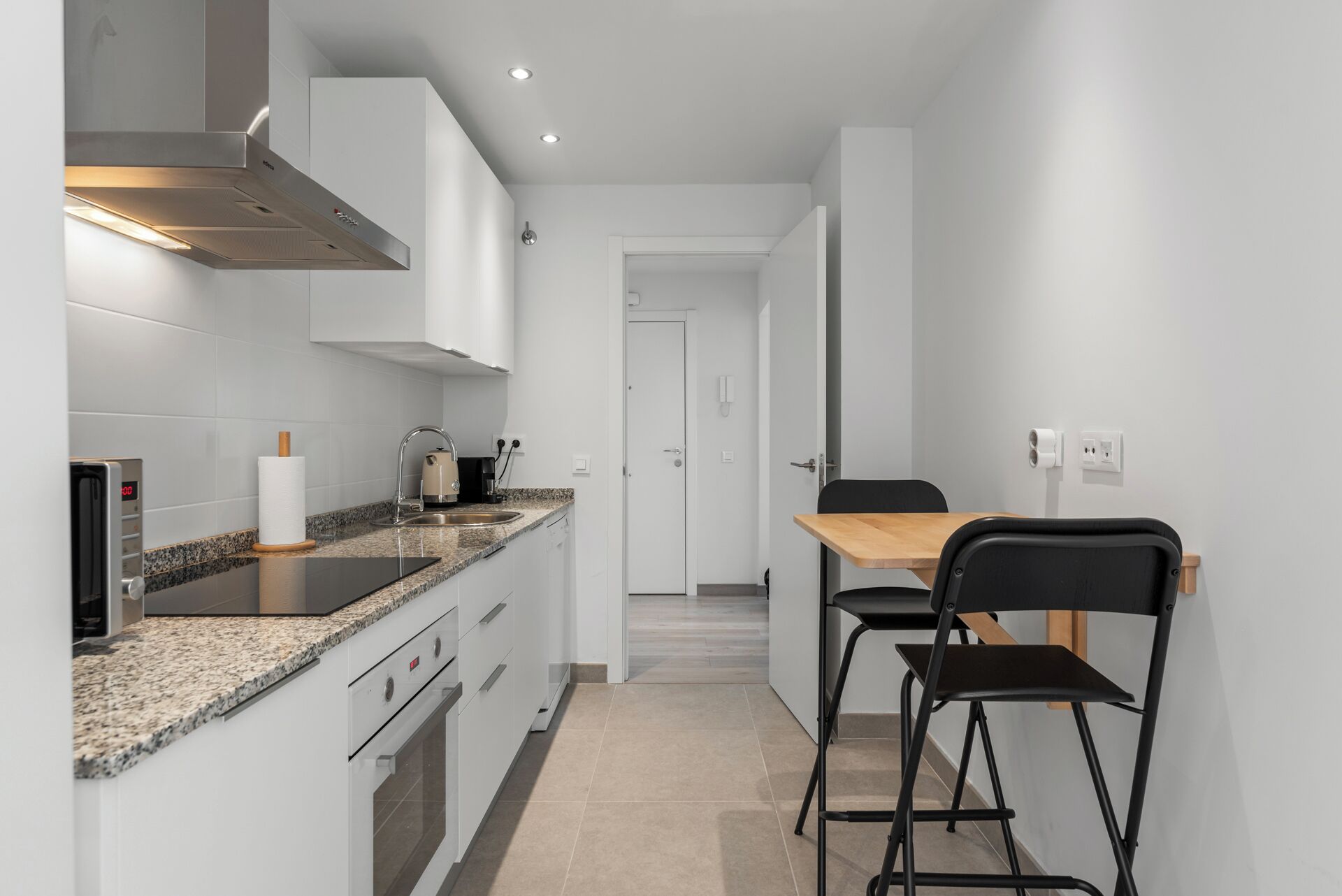 Una cocina moderna con armarios blancos, encimera de granito y campana extractora de acero inoxidable. Hay una zona de comedor con una mesa de madera y sillas negras.