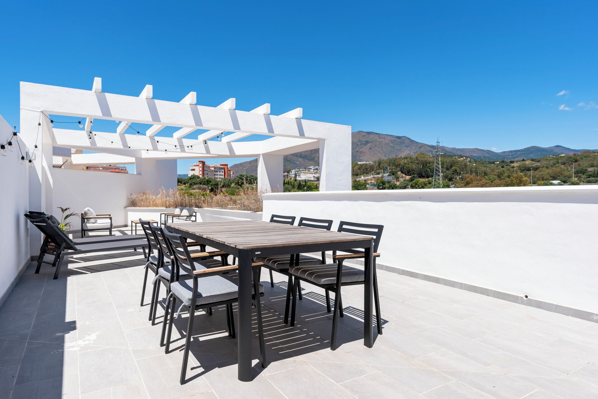 A rooftop terrace with a wooden table and chairs, a pergola, and a view of the surrounding landscape.