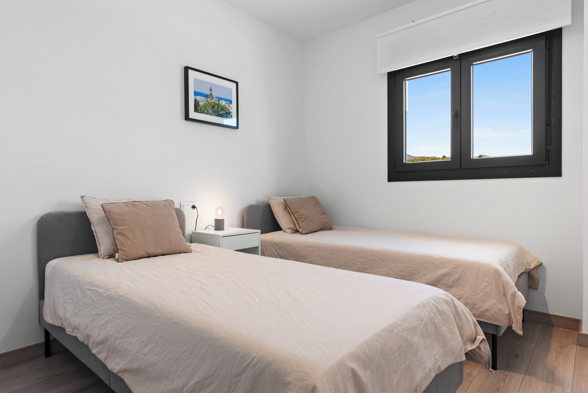 A bedroom with two beds, a nightstand, a bedside lamp, a framed picture on the wall, and a window with a view.