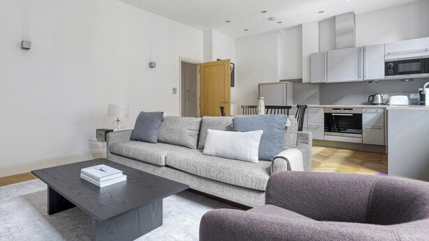 Ein modernes Wohnzimmer mit einem grauen Sofa, einem dunklen Couchtisch und einem violetten Sessel. Im Hintergrund ist eine Küche mit grauen Schränken und einer Mikrowelle erkennbar.