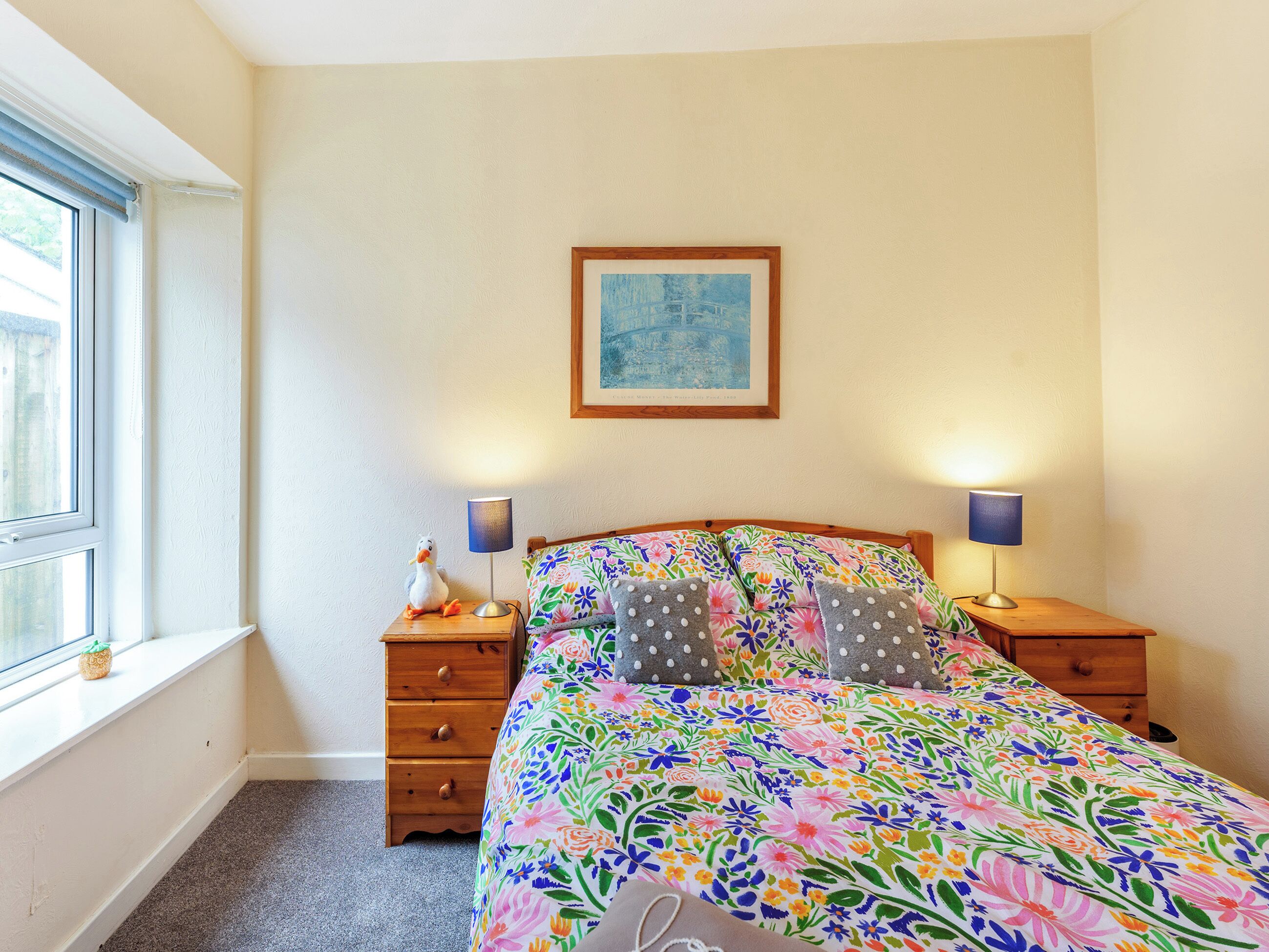 A bedroom with a floral bedspread, two bedside lamps, a wooden nightstand, a small figurine, and a framed picture on the wall.