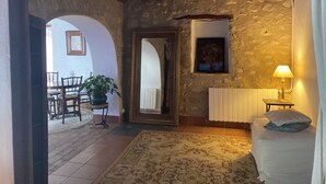 Una habitación rústica con pared de piedra, vigas de madera en el techo, una cama con colchón y almohadas blancas, una mesita con una lámpara y una planta en maceta.