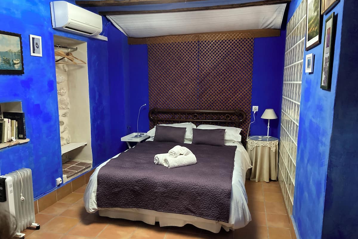 A bedroom with a large bed, a wooden headboard, a nightstand, a wall-mounted air conditioner, a radiator, and a shelf with books.