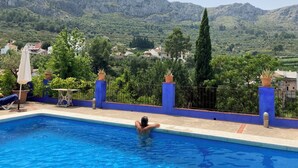Una piscina con una persona en el agua, rodeada por una cerca azul, con vistas a colinas verdes y casas al fondo.