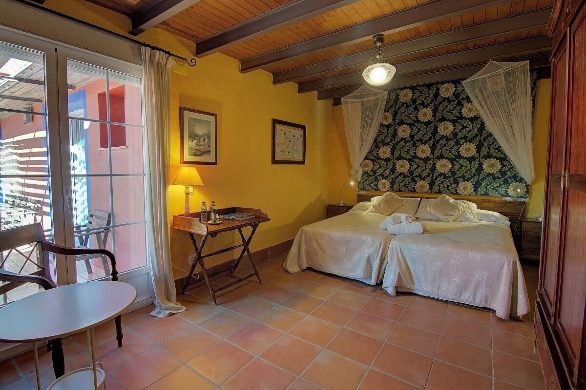 A bedroom with a bed, a table, and a chair. There is a window with curtains, a lamp, and a picture on the wall.