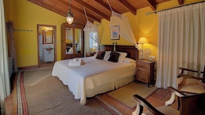 A bedroom with a large bed, wooden ceiling beams, a nightstand with a lamp, and a chair. - Casa Gallinera (Vall de Gallinera)