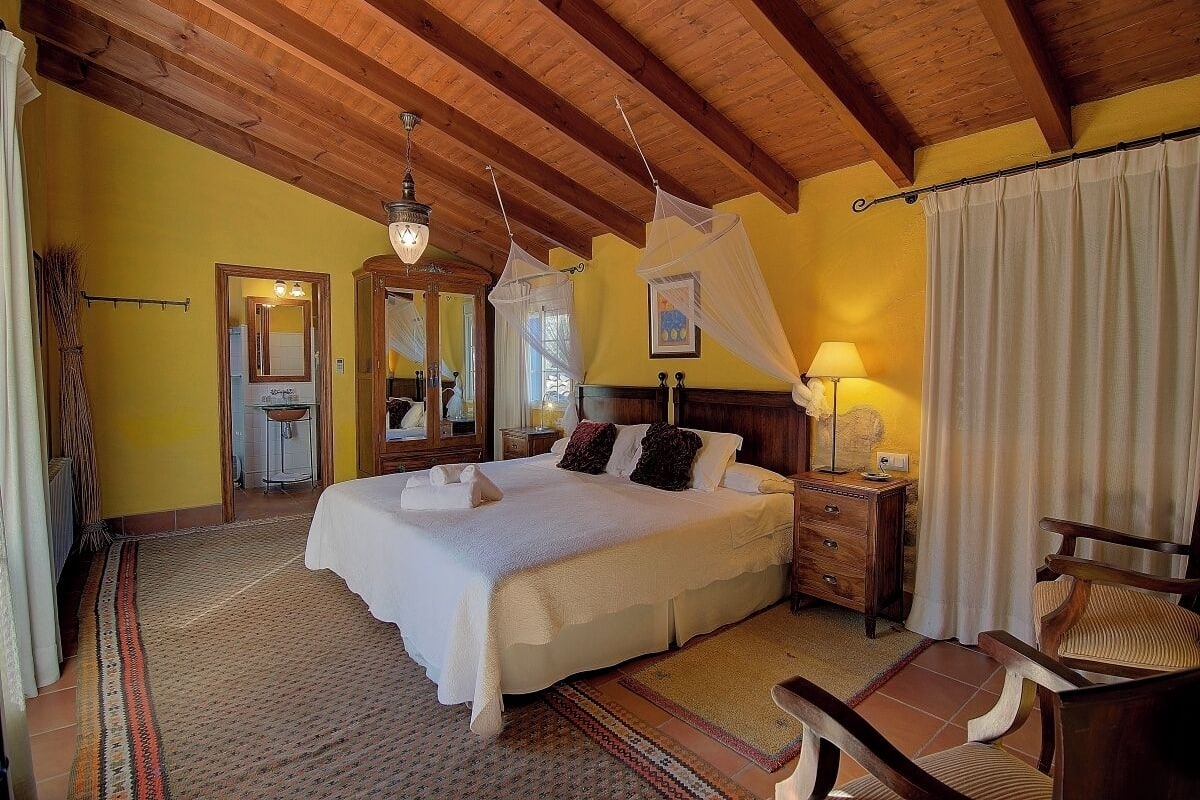 A bedroom with a large bed, wooden ceiling beams, a nightstand with a lamp, and a chair.