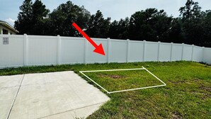 Un jardin avec une clôture blanche, une terrasse en béton et une pelouse délimitée par une ligne blanche.