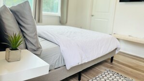 Une chambre à coucher avec un lit, une petite plante en pot, une table de chevet blanche, un téléviseur fixé au mur et une fenêtre avec des rideaux.