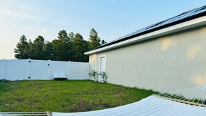 Un hamac et un canapé blanc dans un jardin, avec une clôture blanche, une porte et des arbres en arrière-plan.