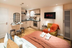 A compact hotel room with a kitchenette, a bed with a pink quilt, a TV displaying 'NETFLIX', and a small seating area with a yellow chair. - 5minwalk2 Bakerstreet/centrallnd (London)