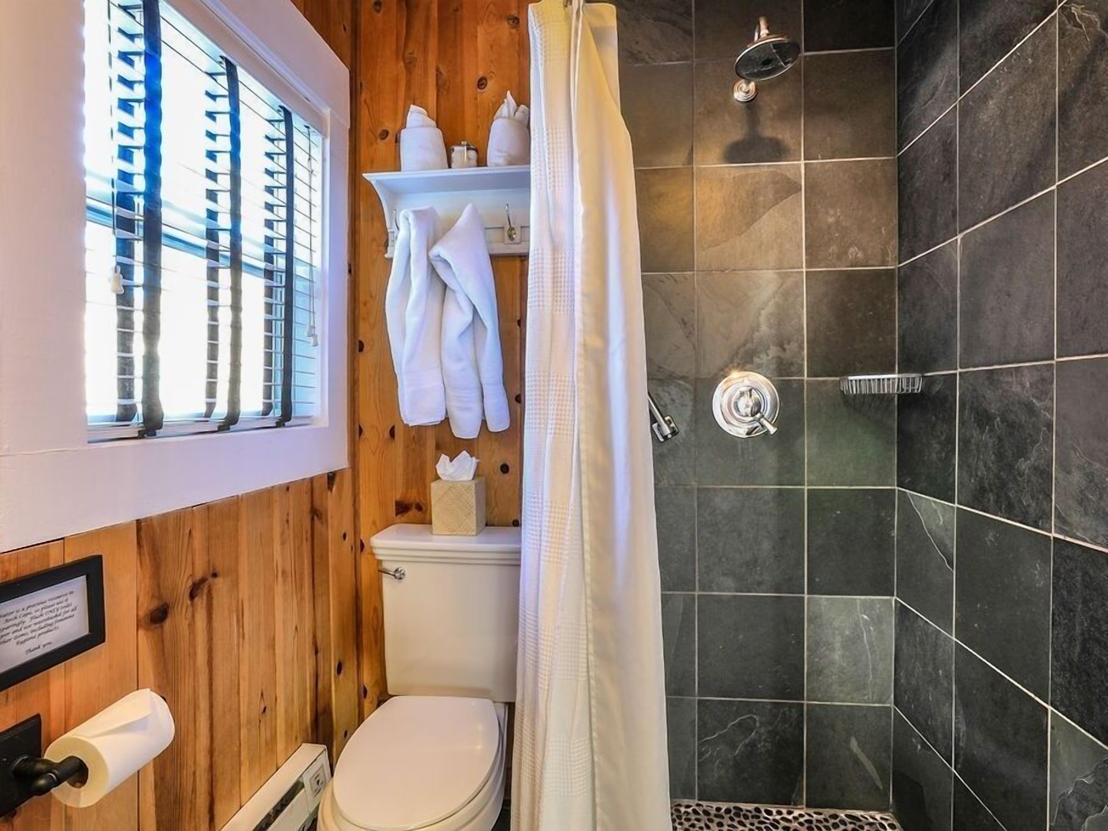 A bathroom with a shower, a toilet, and a window with blinds.