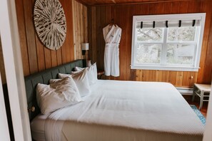 A neatly made bed with white bedding, a round wall decoration, a hanging robe, and a window with blinds.