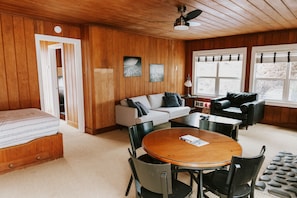 A cozy room with a wooden ceiling, a bed, a sofa, a dining table with chairs, and a window with blinds.