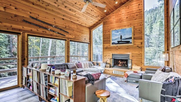 A cozy cabin interior with a fireplace, large windows, and a bookshelf. - Creekside Cabin: Mountain Views, Fireplace & Sauna (Dumont)