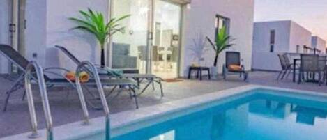 A modern pool area with a clear blue pool, sun loungers, and a glass door leading to a room.