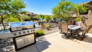 An outdoor patio with a barbecue grill, a table with chairs, and a playground in the background.
