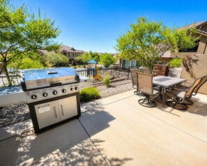 Une terrasse extérieure avec un barbecue, une table et des chaises, et un terrain de jeux en arrière-plan.