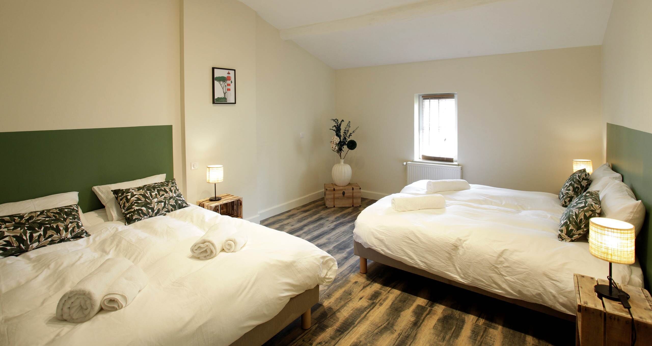 A hotel room with two beds, a window, a vase with flowers, and a lamp on a bedside table.