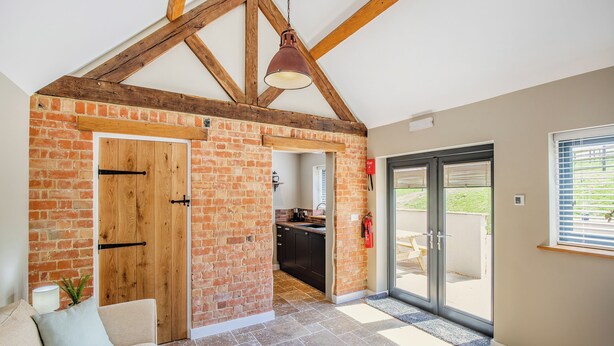 A spacious room with exposed wooden beams, a brick wall, a sliding glass door leading outside, and a cozy seating area with a sofa and cushions. - The Old Stables 2 - UK46683 (Shipton-Under-Wychwood)