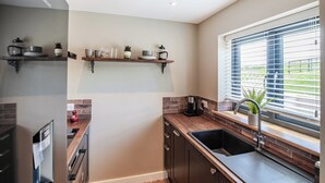 A modern kitchen with a built-in dishwasher, a stainless steel refrigerator, a window with blinds, and a brick backsplash. - The Old Stables 2 - UK46683 (Shipton-Under-Wychwood)