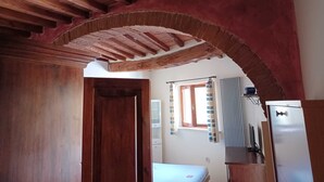 Una stanza con soffitto in legno, un arco in mattoni, un letto, una finestra con tende e un frigorifero.