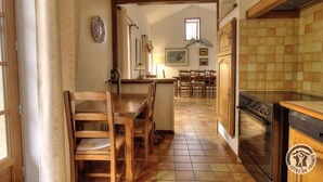 A kitchen with wooden cabinets, a stove, and a dining area with chairs and a table. - La Ponie - Cenves - Gîte (CENVES)