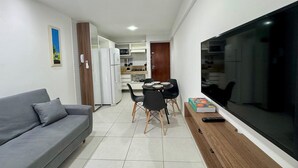 Una sala moderna con un sofá gris, un comedor con mesa y sillas, una cocina con armarios blancos y un televisor grande a la derecha.
