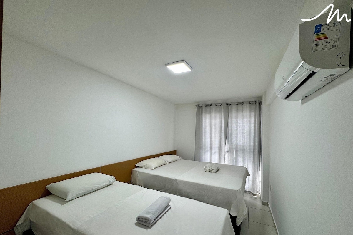 Quarto de hotel com duas camas de solteiro, luminária no teto, ar condicionado e janela com cortinas.