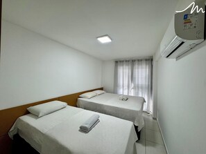 Quarto de hotel com duas camas de solteiro, luminária no teto, ar condicionado e janela com cortinas.