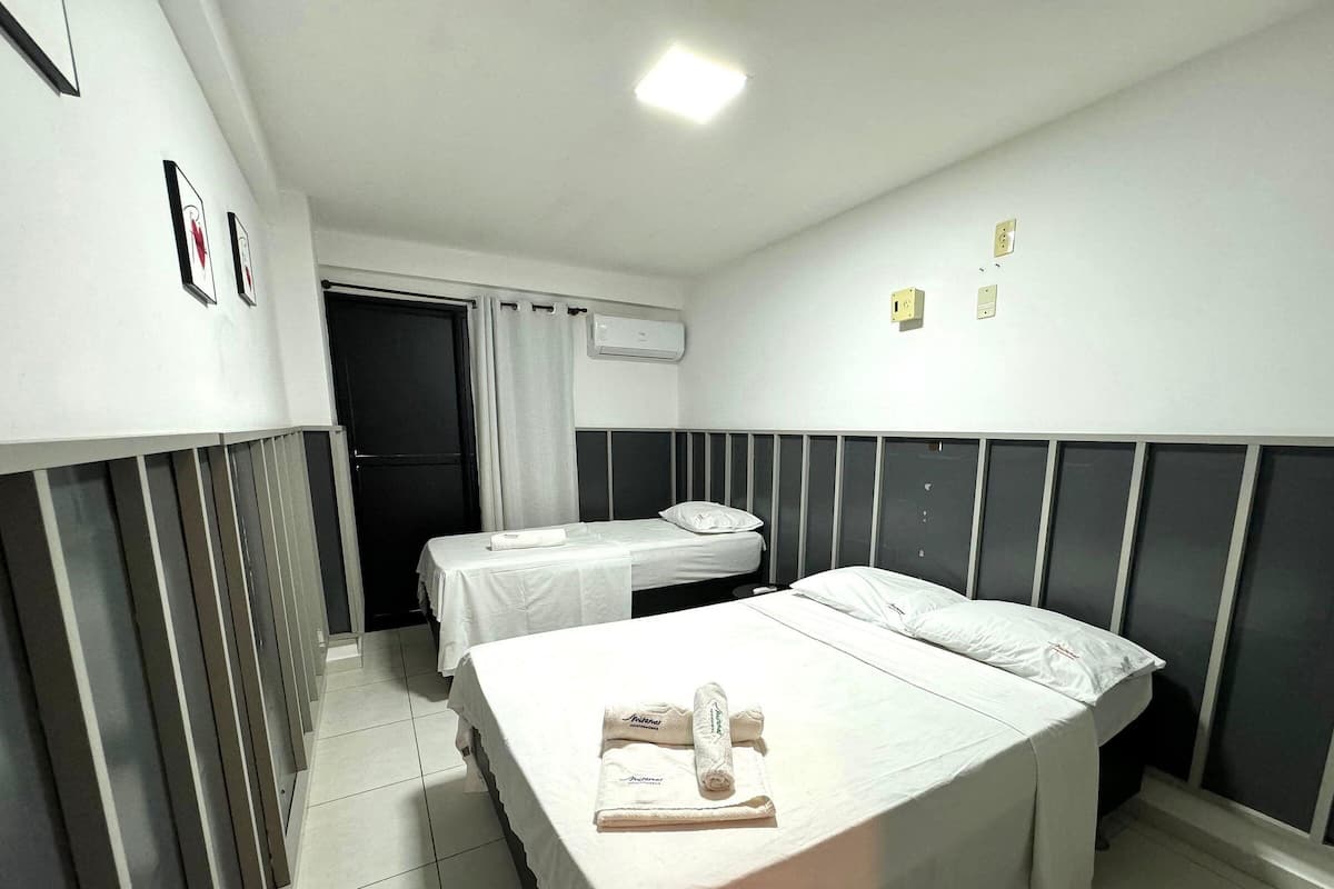 Quarto de hotel com duas camas de solteiro, janela com cortinas, ar condicionado e luminária fixada na parede.
