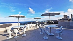 Un espacio al aire libre con sillas y mesas blancas, sombrillas y vistas al océano y al horizonte de la ciudad.