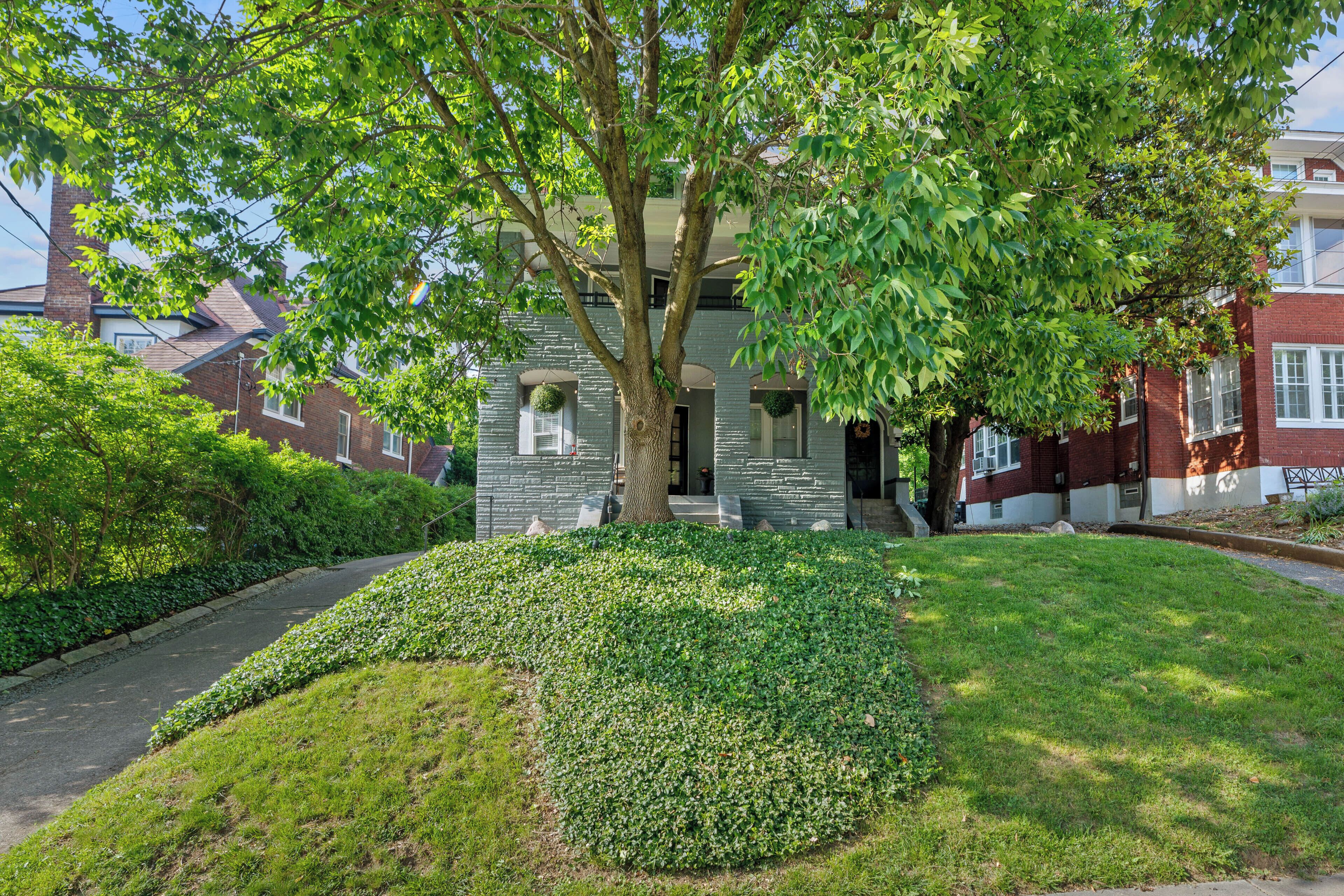 A residential house with a well-maintained lawn, a large tree in front, and a brick facade.