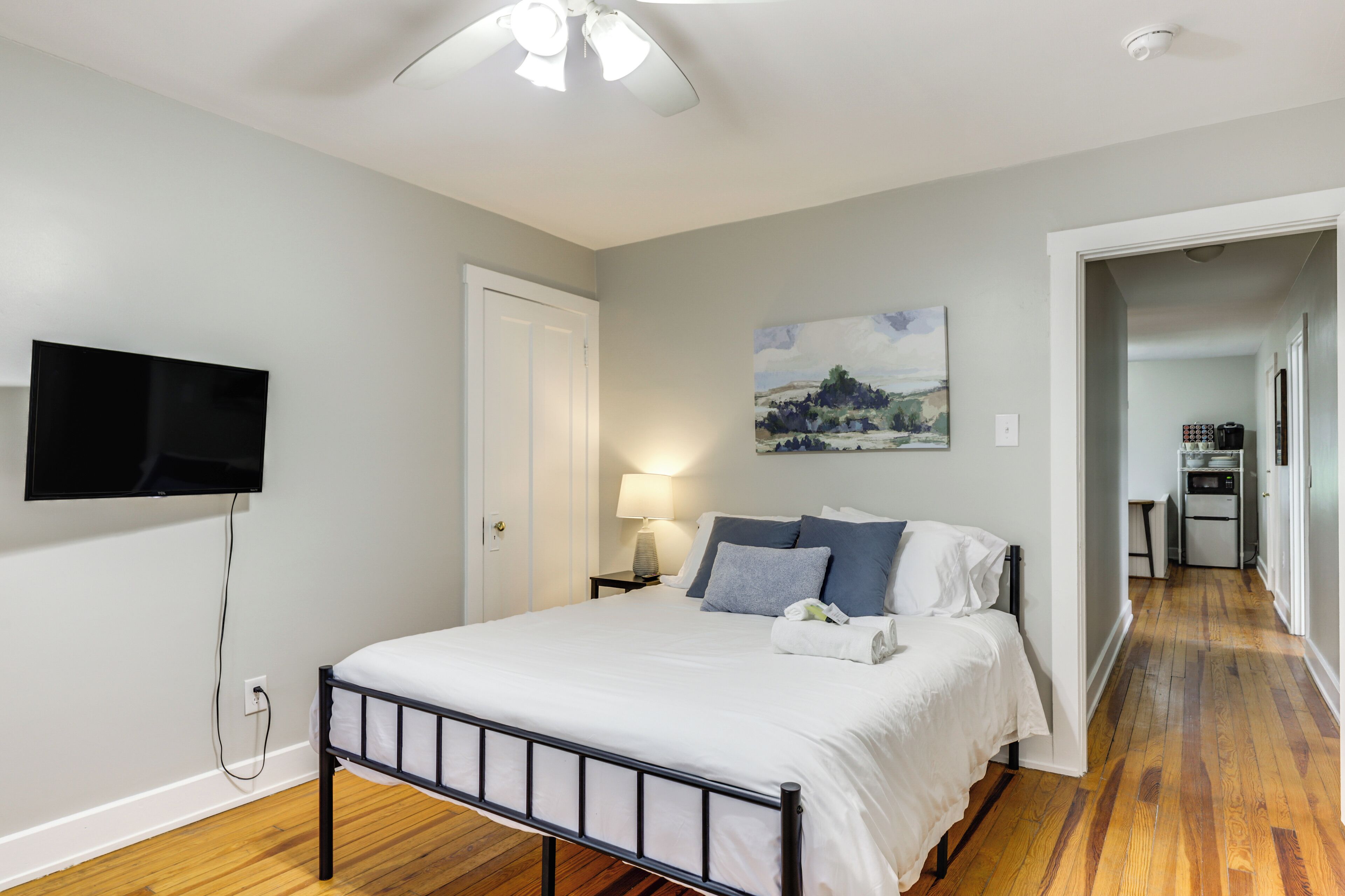 A bedroom with a bed, a TV mounted on the wall, a painting, and a hallway leading to a kitchen.