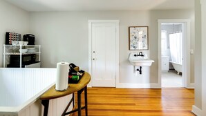Ein Zimmer mit Holzboden, einer weißen Tür, einem weißen Waschbecken, einem gerahmten Kunstwerk und einem Regal mit Küchenutensilien.