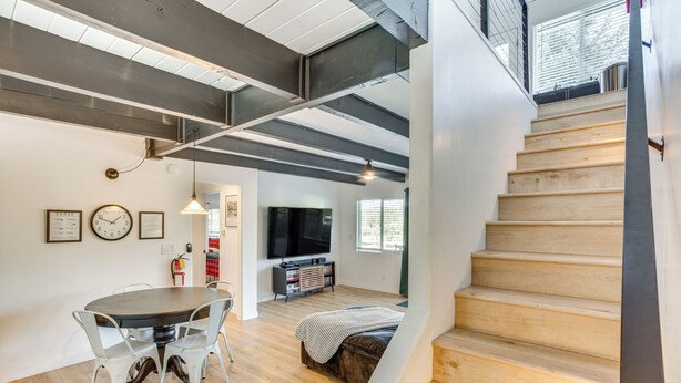 Ein modernes Interieur mit einer Treppe, Holzboden, einem Essbereich mit Tisch und Stühlen, einem Fernseher und einem Bett mit gestreifter Decke.