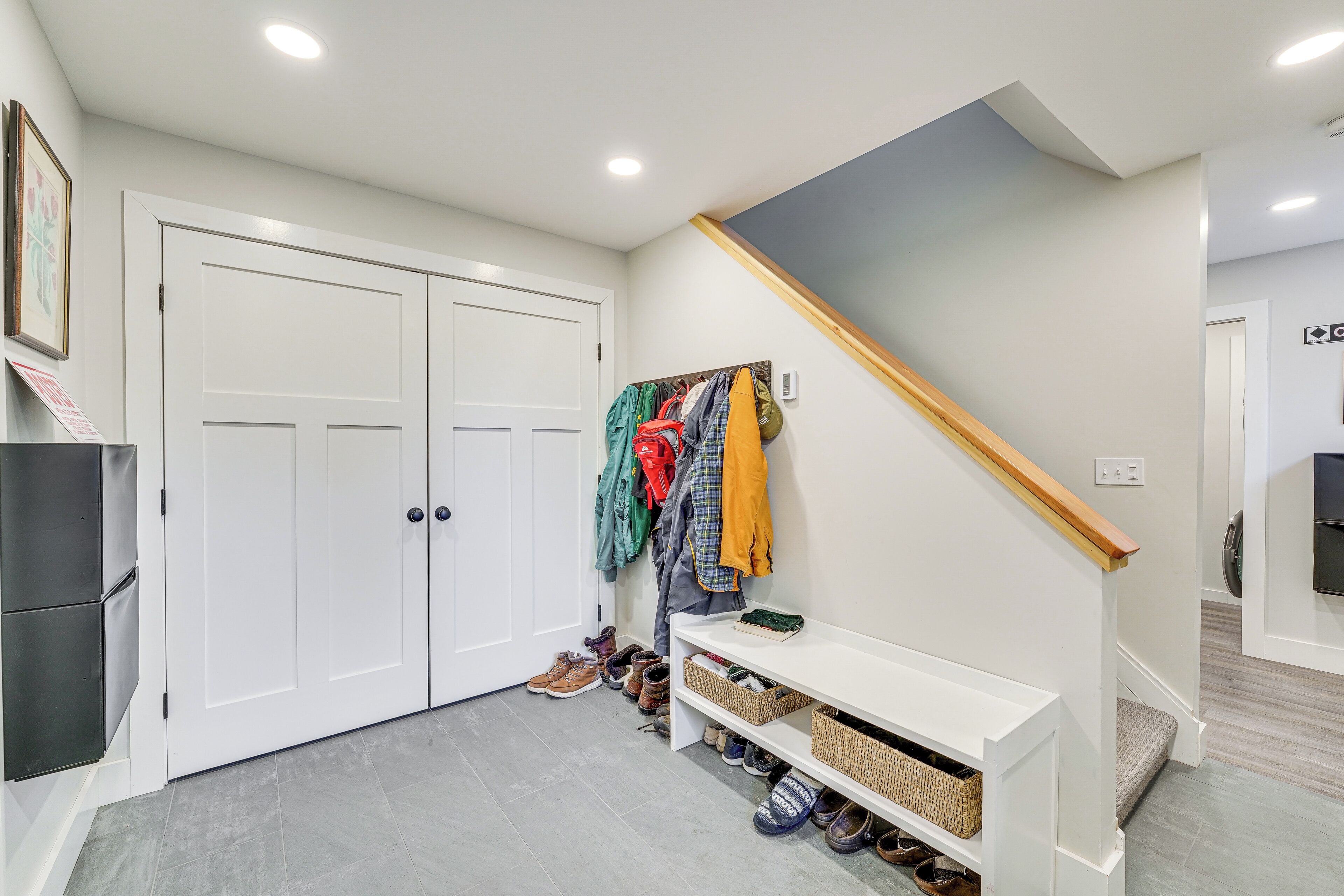 A hallway with a white door, a coat rack with various jackets, a white console table, and a staircase with a wooden handrail.