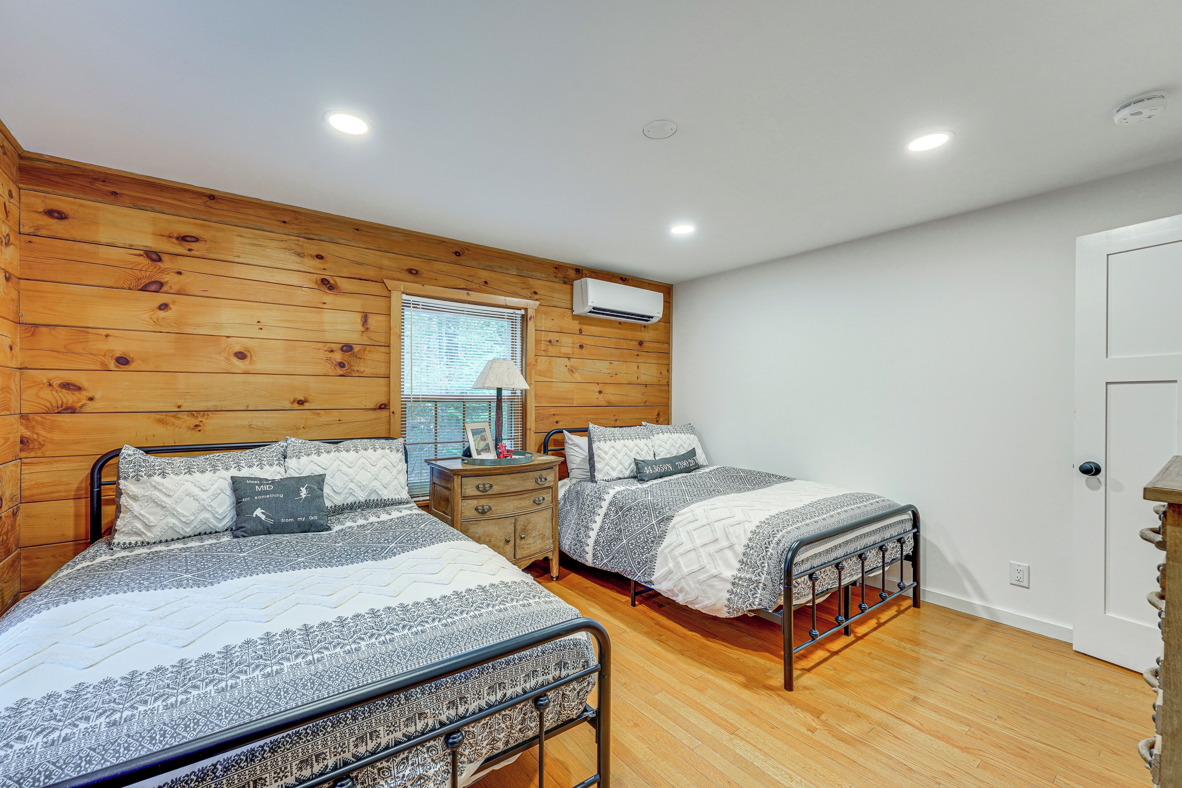 A bedroom with two beds, wooden walls, a window with blinds, an air conditioning unit, and a bedside table with a lamp.