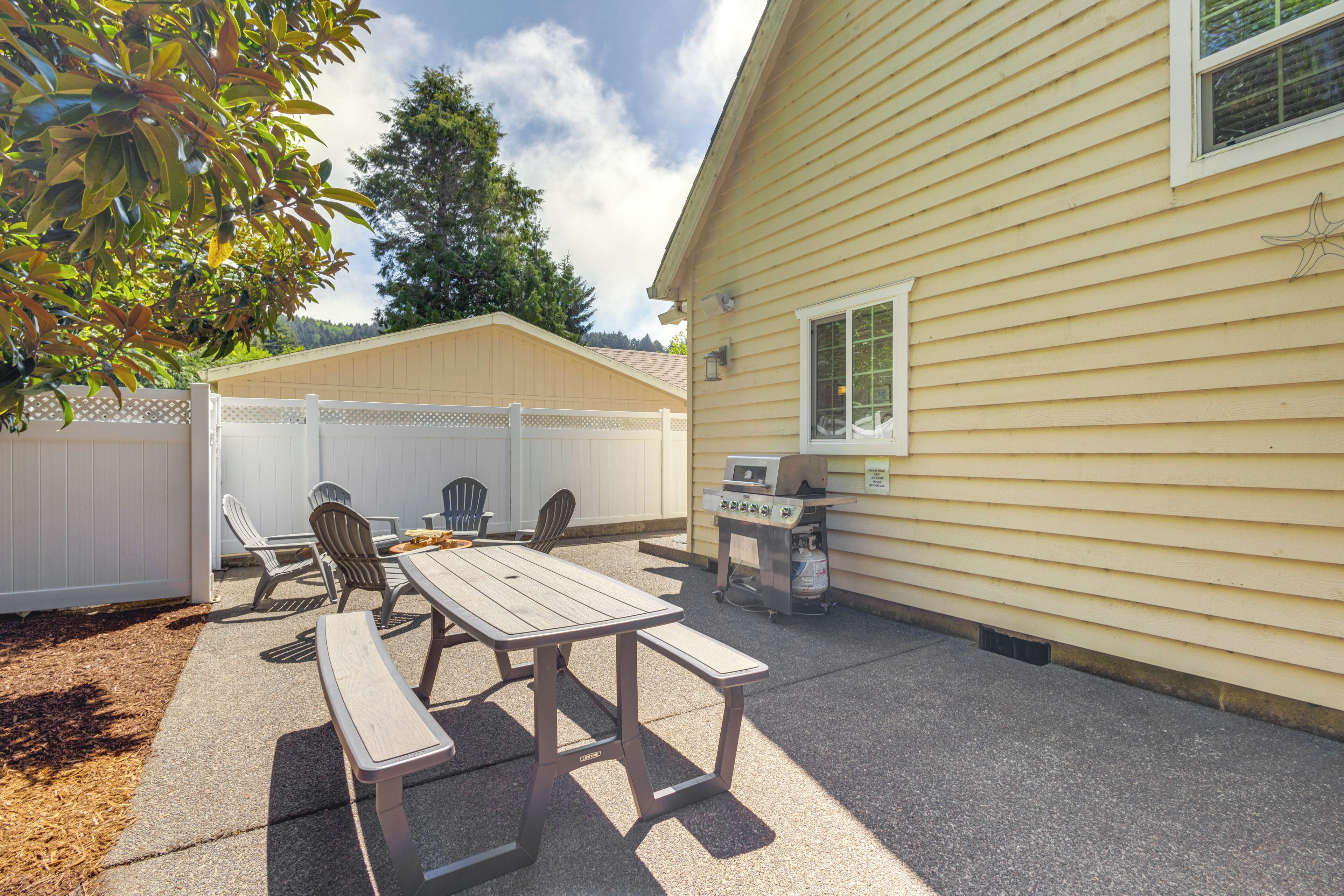 An outdoor patio with a wooden table and chairs, a grill, and a white fence.