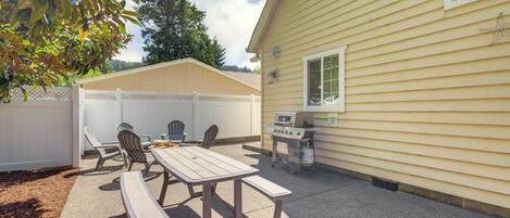 An outdoor patio with a wooden table and chairs, a grill, and a white fence.