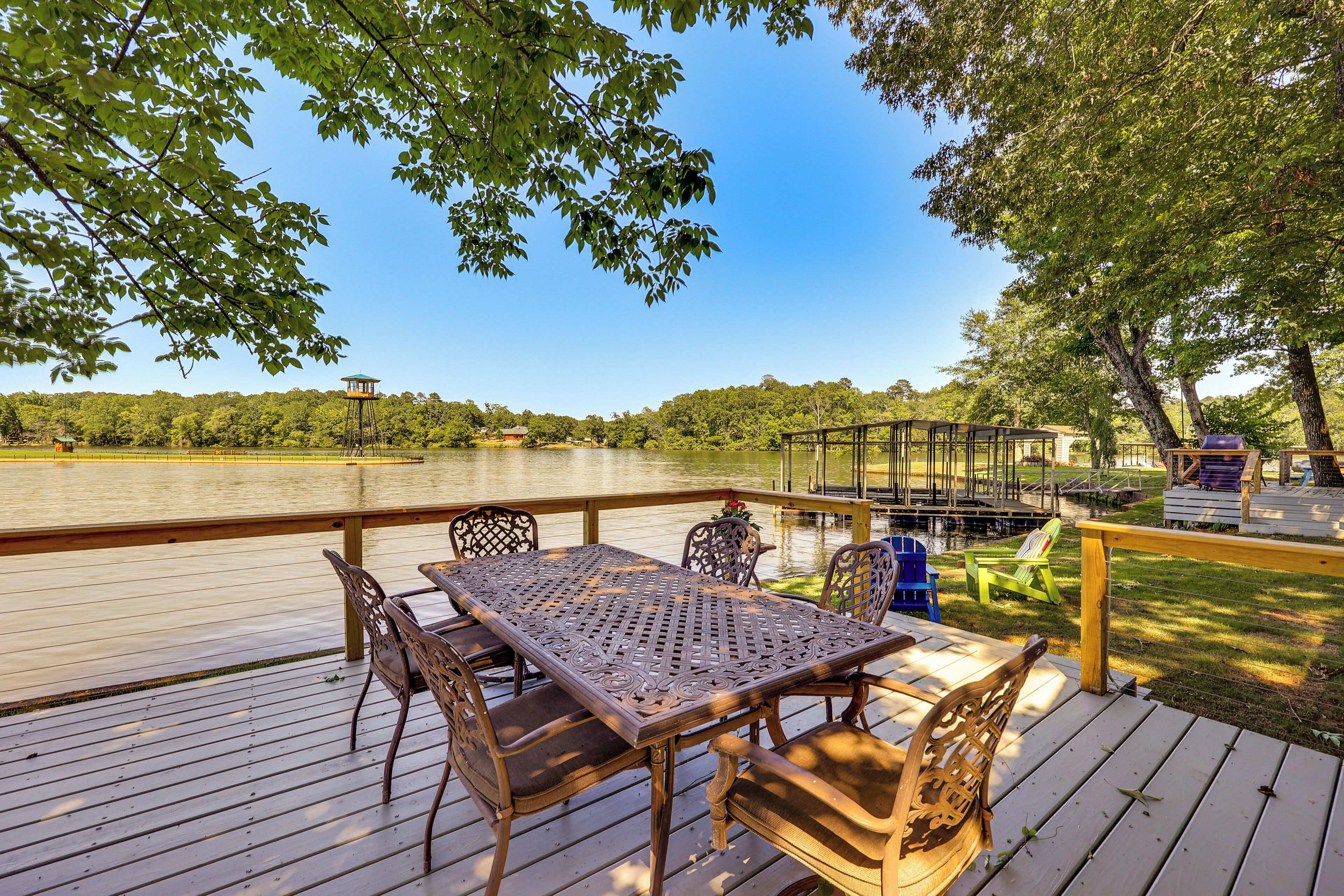 Een houten terras met tafel en stoelen met uitzicht op een meer, omringd door bomen en een houten hek.