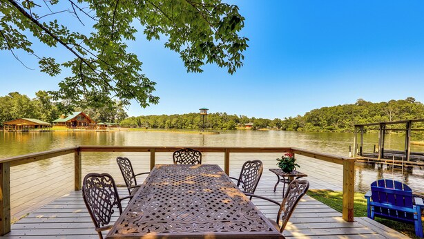 A wooden deck with a table and chairs overlooking a lake, surrounded by trees and a distant building. - Waterfront Lake Hamilton Vacation Rental w/ Dock! (Royal)