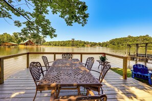A wooden deck with a table and chairs overlooking a lake, surrounded by trees and a distant building. - Waterfront Lake Hamilton Vacation Rental w/ Dock! (Royal)