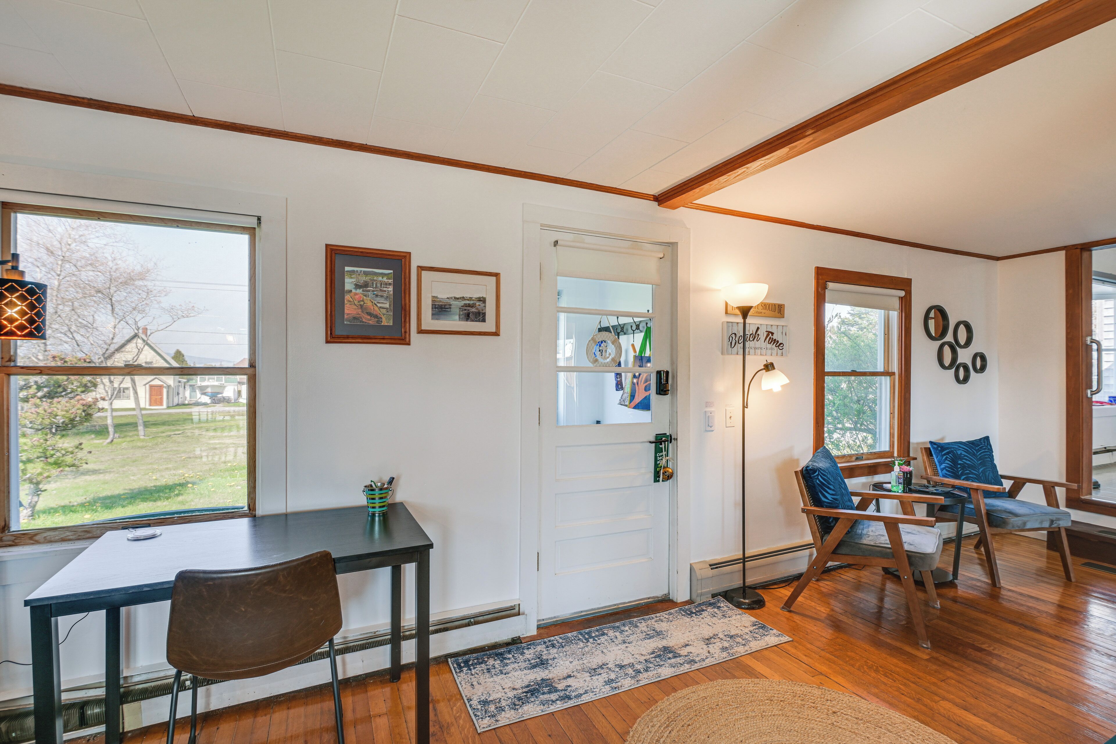 A room with a wooden floor, a white door, a table with a chair, and a window with a view of a yard.