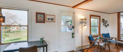 A room with a wooden floor, a white door, a table with a chair, and a window with a view of a yard.