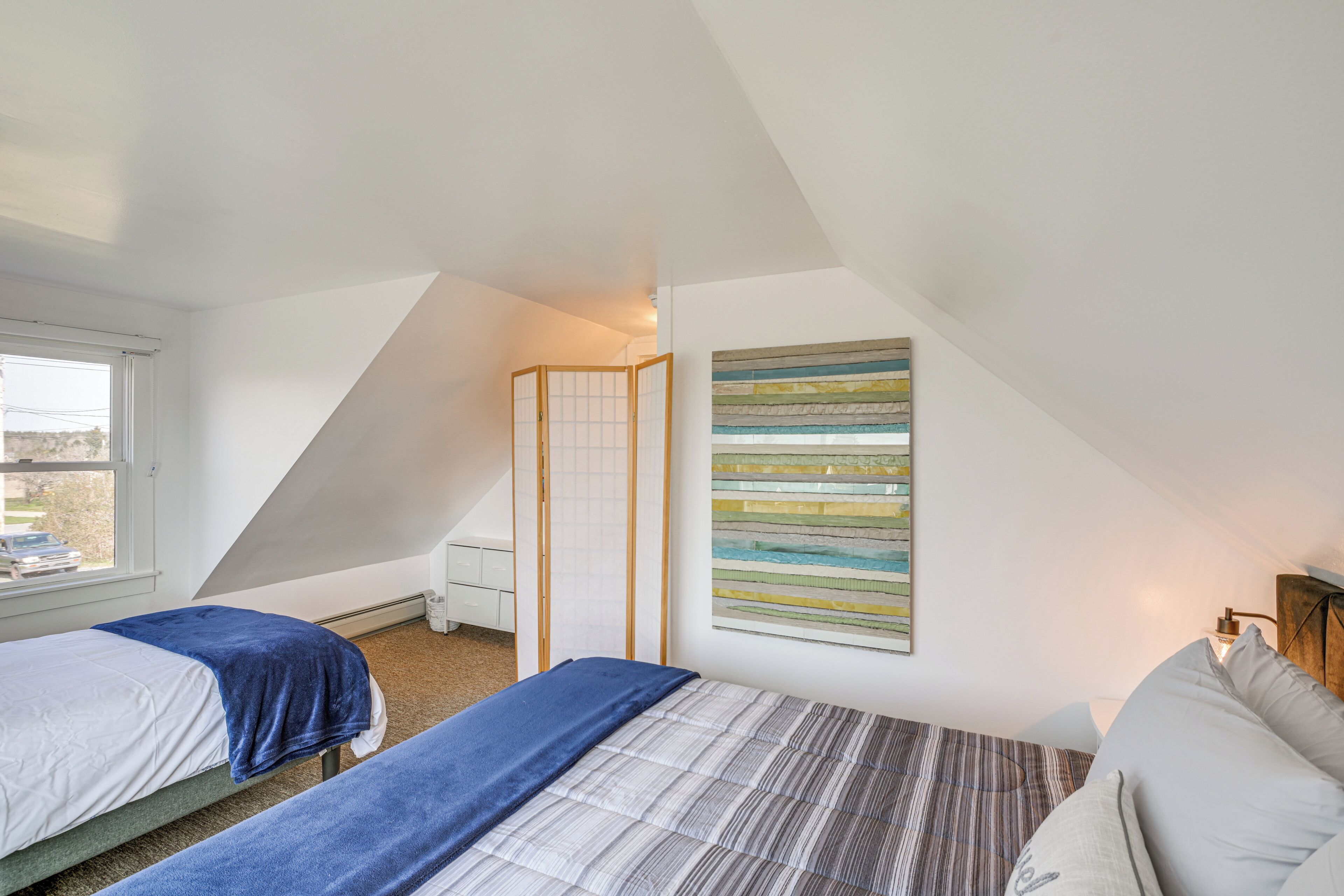 A bedroom with two beds, a window, a painting, and a partition.