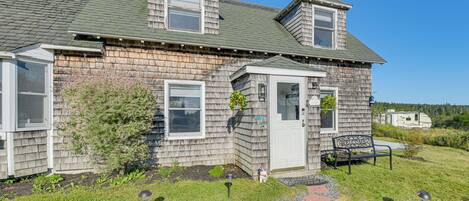 A two-story house with a sloped roof, a front porch, and a small garden area with a bench.